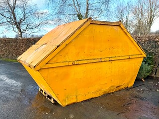 Large yellow industrial skip container standing on wet asphalt in an outdoor area. The metal waste container is shown from the side with a closed top, displaying strong geometric shape and bold color 