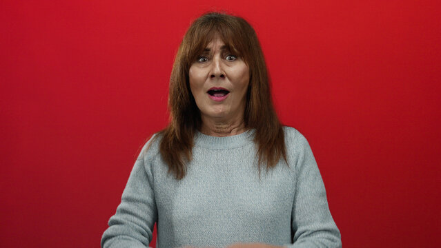 Hispanic senior woman with wide eyes covers mouth in surprise against a bold red wall wearing a gray sweater evoking emotions of shock and disbelief in a stark contrast setting.