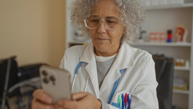 Woman doctor with grey hair using smartphone in hospital clinic room wearing white coat and glasses focused on technology surrounded by medical supplies and stethoscope.