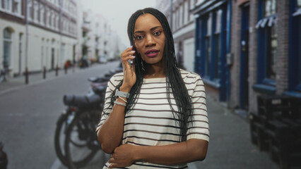 Fototapeta premium Woman holding smartphone, looking at screen with hand visible, wearing striped top and bracelets on a narrow city street near bicycles and storefronts; quiet concern.