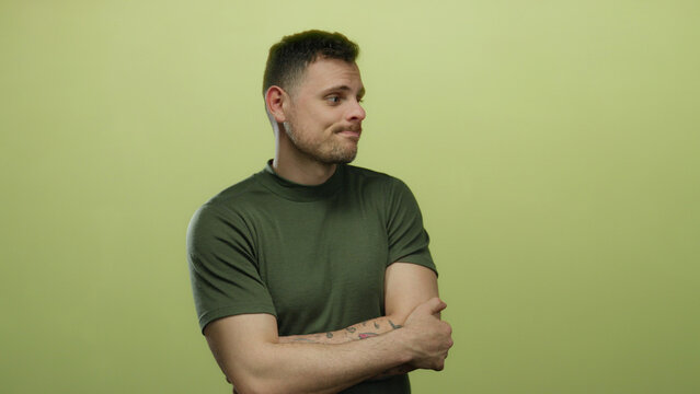Young man with beard and tattoos stands isolated against a yellow background expressing indecision and contemplation while wearing a casual green shirt.