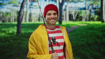 Naklejka premium Young man smiling in a vibrant park setting, wearing a red beanie and yellow jacket, holding a fishing rod with lush green trees in the background.