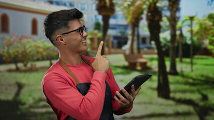 Fototapeta premium Young man smiling outdoors holding tablet in park setting, wearing glasses and apron, conveying attractive, casual, relaxed, urban vibe in sunlit exterior.