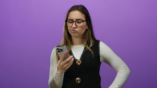 Young woman wearing glasses looks puzzled while holding smartphone against isolated purple background suggesting technology misunderstanding or confusion moment