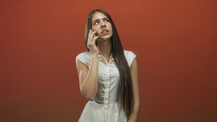 Fototapeta premium Woman holds smartphone to ear with hand, wearing white dress in studio against orange backdrop; pensive.