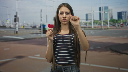 Fototapeta premium Teen woman holds a small red heart in one hand while raising a closed fist toward the camera on a city street; heartbreak longing.