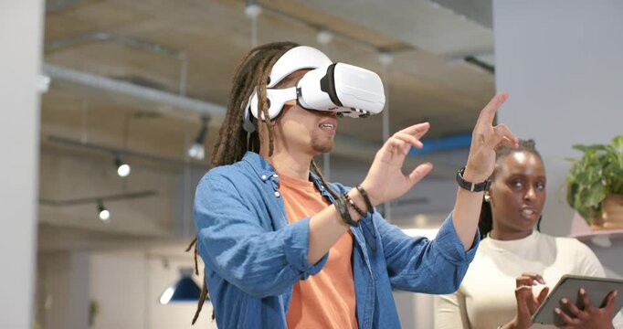 African American staff testing VR in office, headset user using controls while colleague on tablet