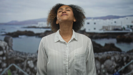 Woman tilts head back showing neck at seaside town by rocky shore under cloudy sky now; serenity...