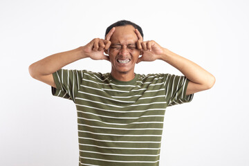 An Asian man wearing a green shirt, standing against a plain white background, holding his head with hands and expressing a painful or stressed facial expression, depicting a headache