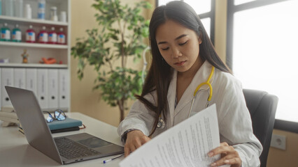 Young woman doctor with stethoscope working on a laptop in a clinic room filled with files and...