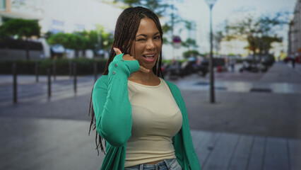 Fototapeta premium Young african american woman smiling and showing call gesture with hand and wink on a busy urban street; playful confidence.