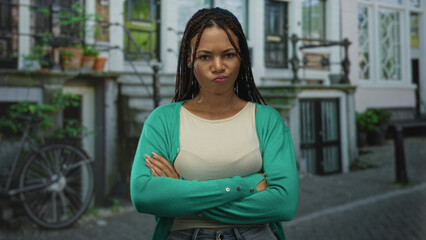 Woman with crossed arms and pursed lips on a city street in front of townhouse building wearing...