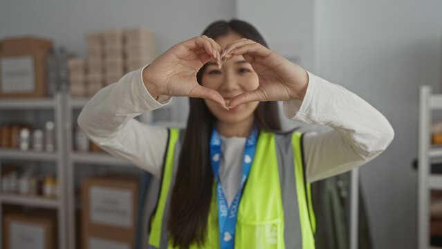 Young woman in a reflective vest makes a heart gesture at a volunteer center, surrounded by charity supplies, emphasizing community and kindness.