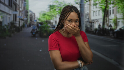 Woman covering mouth with hand on a city street wearing red shirt and bracelets, smiling with...
