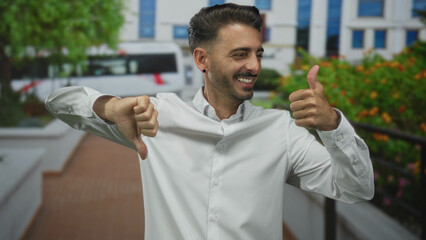Young hispanic man in white shirt gives thumbs up and thumbs down gesture on busy street outdoors;...