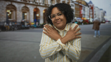 Fototapeta premium Young african american woman wearing glasses crosses arms on street lined with stone buildings; anxiety.