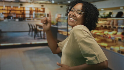 African american woman smiling and dancing with pointing finger in a bakery building; youthful joy...
