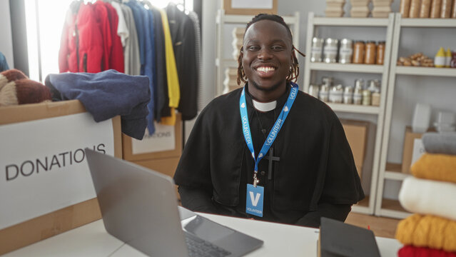 Man volunteer smiling at camera, wearing clerical collar and volunteer lanyard, seated with laptop and donation boxes in a building; compassion service community.