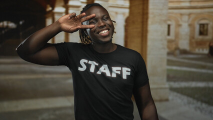 Man wearing staff tshirt shows peace sign to eye on a cobbled street with stone arches in old town; joy confidence.