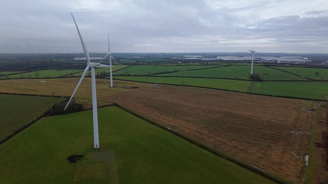 Aerial view of wind turbines standing tall amidst the patchwork of green and golden fields, creating a striking contrast against the overcast sky, Yelvertoft, Crick, United Kingdom.