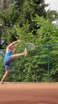 Male Tennis Player On Clay Performing Dramatic Airborne Serve With Racket Raised, misses the ball, tries to jump to hit the ball. Blue Outfit, Dusty Orange Court, Energetic Midair Motion. Slow motion