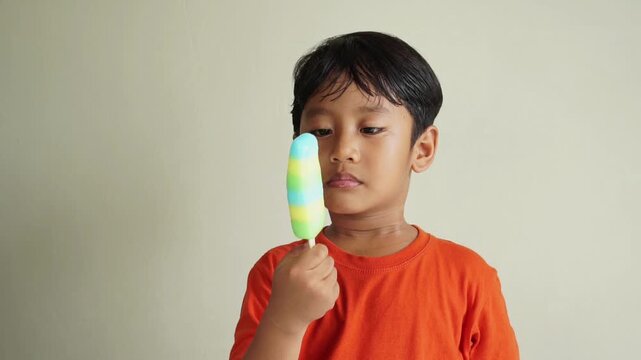 Close-up of a young boy in an orange t shirt enjoying a colorful popsicle on a light background
