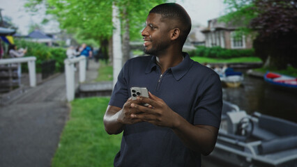 Man holding smartphone with both hands and smiling while looking at screen on street beside canal boats and pedestrian walkway; contentment.
