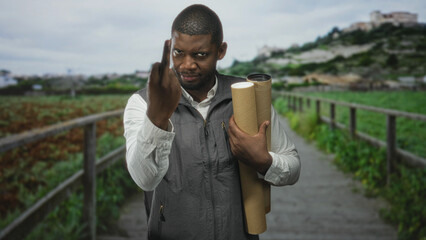 Man architect holding rolled blueprints and coffee cup shows middle finger toward camera on street near houses and railing; defiance.