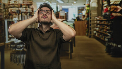 Obraz premium Man covering ears with hands in building clothing store aisle, glasses visible, bare forearms and hands prominent; frustration overwhelm.