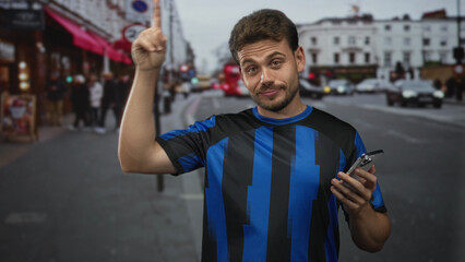 Man pointing at smartphone on street, smiling and holding phone in right hand while wearing a blue striped jersey; joy connection.