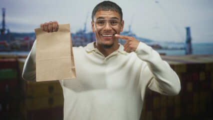 Young man smiling while holding a paper bag and pointing finger to cheek on a street by a shipping port with stacked containers; pride.