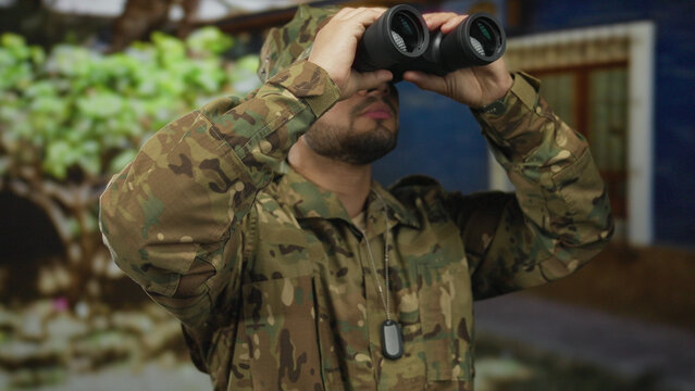 Man in camouflage uniform holding binoculars and looking upward by green foliage near building; duty vigilance.