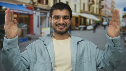 Naklejka premium Man in light blue jacket holds arms wide under storefront awnings in sunlit bustling city street; confidence.