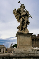Obraz premium Stone angel statue on a historic bridge in Rome, Italy, photographed from a low angle against a dramatic sky, with St. Peter’s Basilica visible in the background