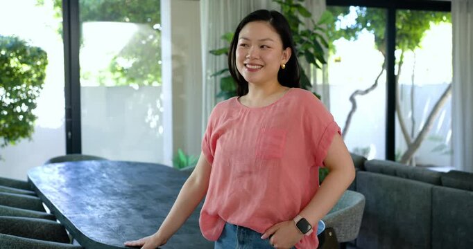 Adult Asian female leaning on dark dining table in home, camera moving closer, smiling to engage