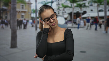 Fototapeta premium Woman wearing glasses and black top with hand on cheek, resting head on hand on a busy city street; tired solitude.