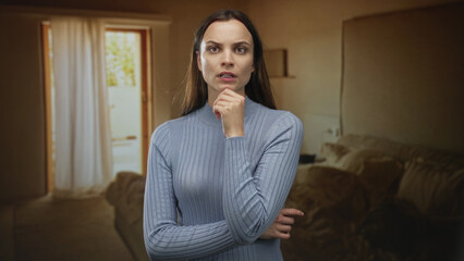 Obraz premium Woman with hand on chin and folded arms in a building living room near doorway, looking to side; thinking doubt.