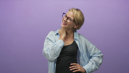 Fototapeta premium Woman wearing glasses places right hand on neck and left hand on hip in studio portrait against purple seamless backdrop; contentment.