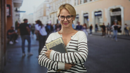 Obraz premium Woman holding ornate hardcover book, arms crossed and smiling, wearing striped sweater and glasses on a busy street; confidence study.