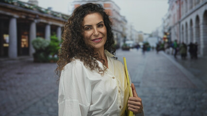 Woman holding yellow folder and leather bound journal while smiling on cobblestone city street sidewalk; optimism ambition.