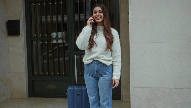 Young woman talking on phone with suitcase on street, wearing casual sweater and jeans, standing outdoors near entrance on a city sidewalk.