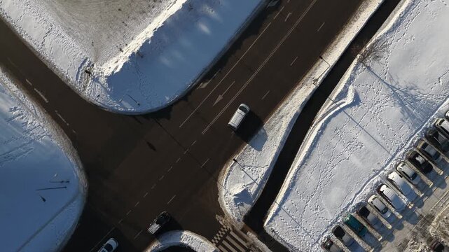 Top-down drone footage of cars driving on parallel roads in winter, surrounded by snow-covered ground, trees, and pedestrian paths under low winter sunlight.