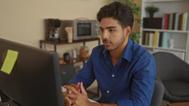 Man with folded hands at desk wearing blue shirt, watching a computer monitor in studio; contemplative.