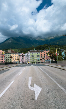 View of a long road with a directional arrow leading towards colorful buildings nestled against imposing mountains under a dramatic sky, Innsbruck, Tyrol, Austria.