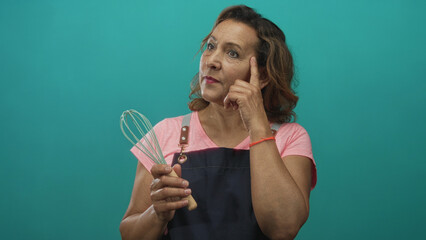 Fototapeta premium Middle aged hispanic woman in apron holds metal whisk and points finger upward in studio; inspiration creativity motivation idea.