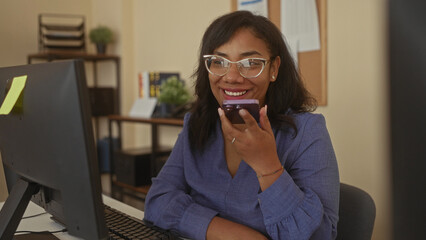 African american woman holding smartphone to mouth at office desk with computer monitor, smiling...