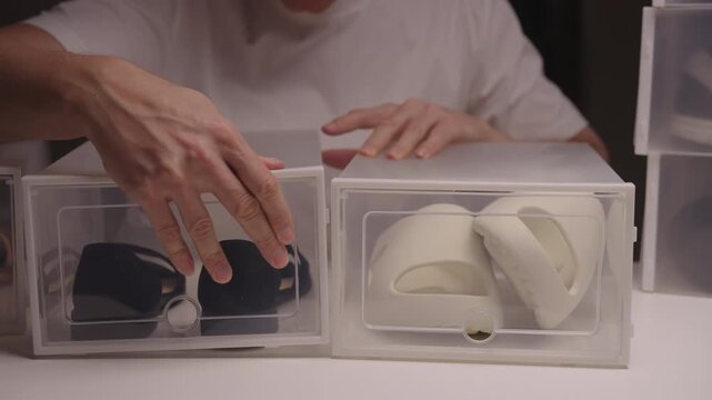 Close-up front view of young man organizing and storing seasonal shoes in clear plastic containers, keeping neatly arranged and tidy, promoting home clean-up and decluttering efforts, slow motion.