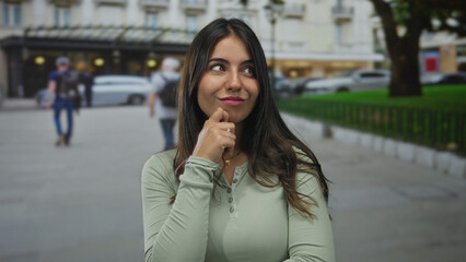 Obraz premium Hispanic woman contemplating outdoors on a bustling city street with blurred pedestrians in the background, showcasing thoughtful expression amidst urban life.