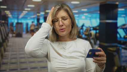 Fototapeta premium Woman holding blue creditcard and hand to head in gym building with eyes closed and rows of treadmills visible; financial stress.