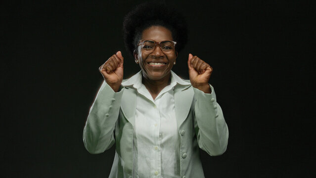 Woman smiling confidently over isolated black background wearing glasses and green suit representing positivity and joy.